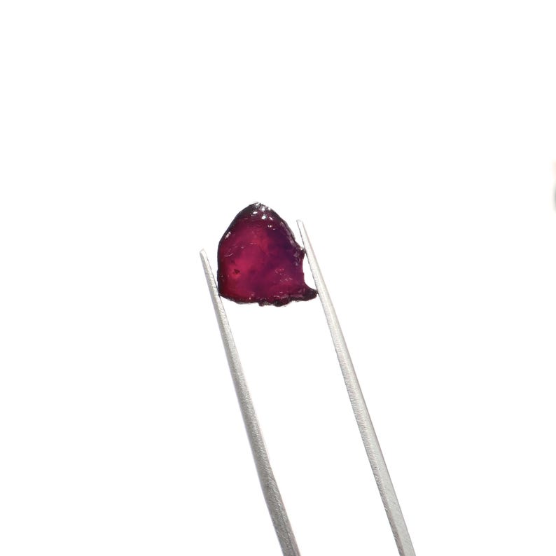 May include: A raw, uncut gemstone held by silver tweezers. The gem is a deep, translucent red-purple color with a rough, irregular shape. The background is plain white, highlighting the gem's natural beauty.