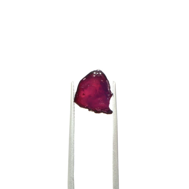 May include: A small, irregularly shaped, translucent gemstone with a deep red hue, held by silver tweezers against a white background. The gem's surface reflects light, showcasing its color variations.