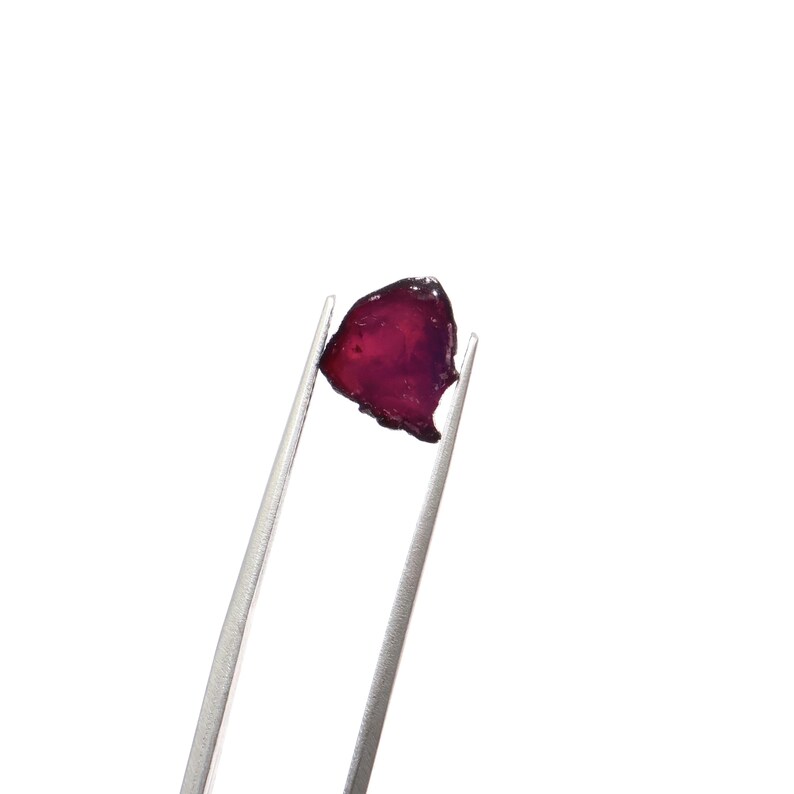 May include: A small, irregularly shaped, deep red gemstone is held by silver tweezers against a white background. The gem has a translucent quality, with variations in color from dark red to a purplish hue. The edges of the stone are rough and uneven.