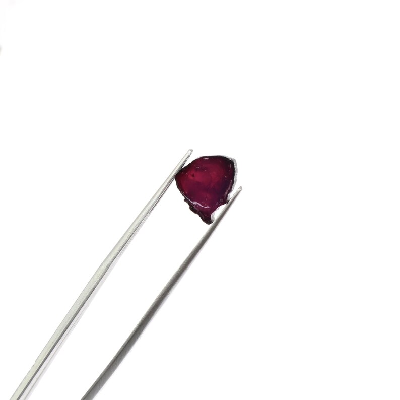 May include: A deep red, irregularly shaped gemstone is held by a pair of silver tweezers against a white background. The gem has a translucent quality, with a slightly uneven surface. The tweezers are positioned diagonally, showcasing the gem's unique form.
