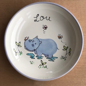 Pode incluir: Uma tigela de cerâmica branca com uma borda azul, apresentando um hipopótamo de desenho animado com borboletas e plantas verdes. A tigela é personalizada com o nome "Lou".