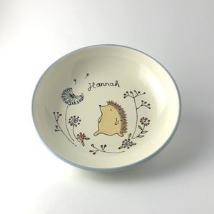 May include: A white ceramic bowl with a blue rim, featuring a hand-painted design of a hedgehog walking through a field of flowers. The bowl is personalized with the name "Hannah".