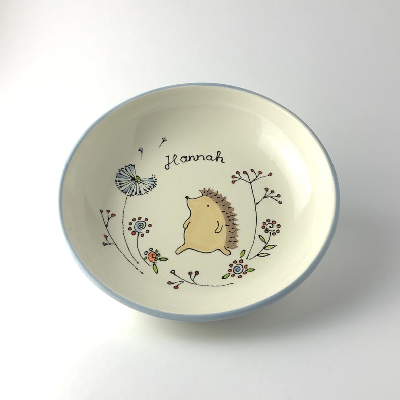 May include: A white ceramic bowl with a blue rim, featuring a hand-painted design of a hedgehog walking through a field of flowers. The bowl is personalized with the name "Hannah".