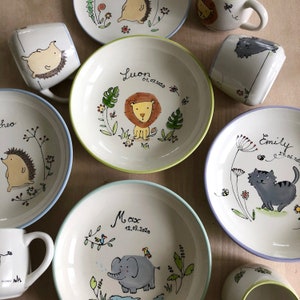May include: Set of personalized ceramic bowls and mugs with animal illustrations. The bowls feature a lion, elephant, and cat, while the mugs have a hedgehog and a cat. Each piece is decorated with a name and date.