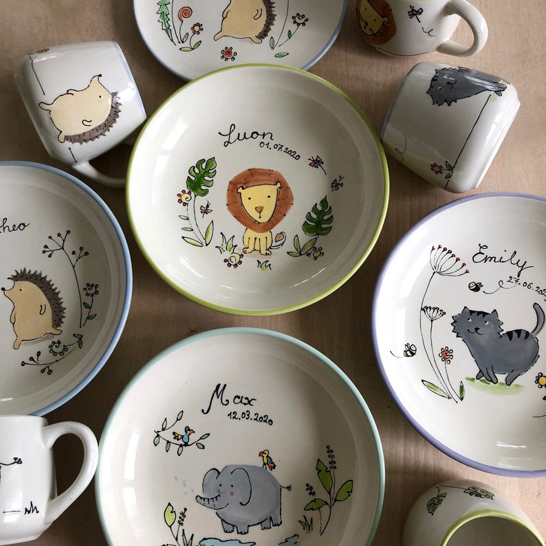 May include: Set of personalized ceramic bowls and mugs with animal illustrations. The bowls feature a lion, elephant, and cat, while the mugs have a hedgehog and a cat. Each piece is decorated with a name and date.