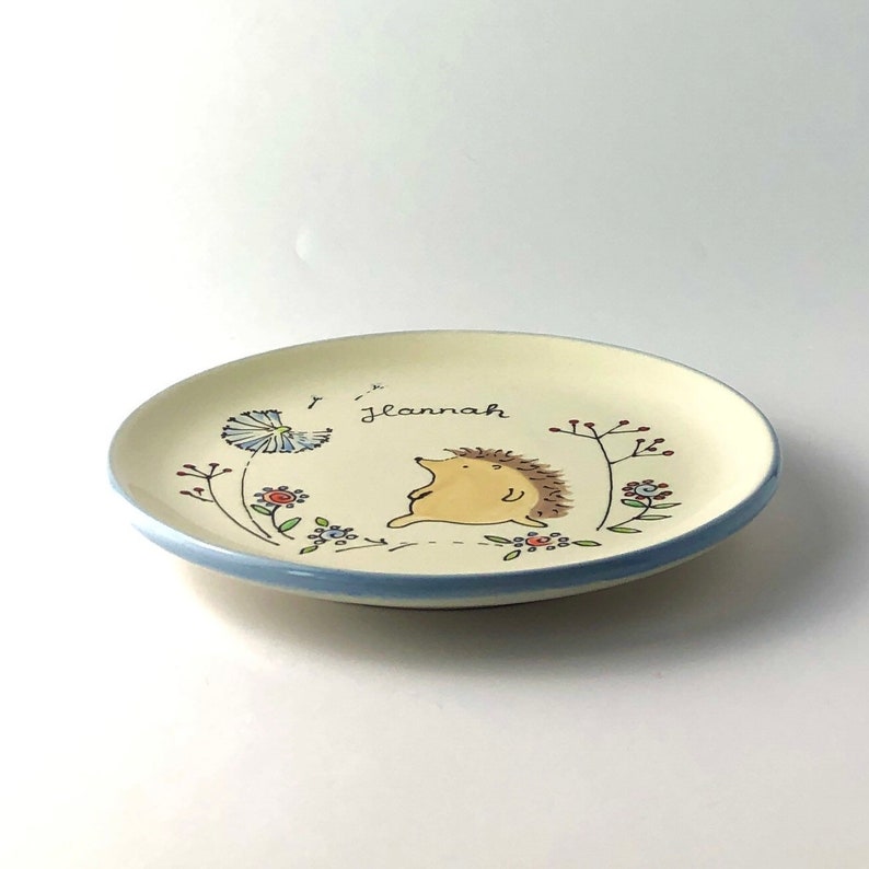 May include: A white ceramic plate with a blue rim features a hand-painted design of a hedgehog, flowers, and a dandelion. The plate is personalized with the name "Giannah".