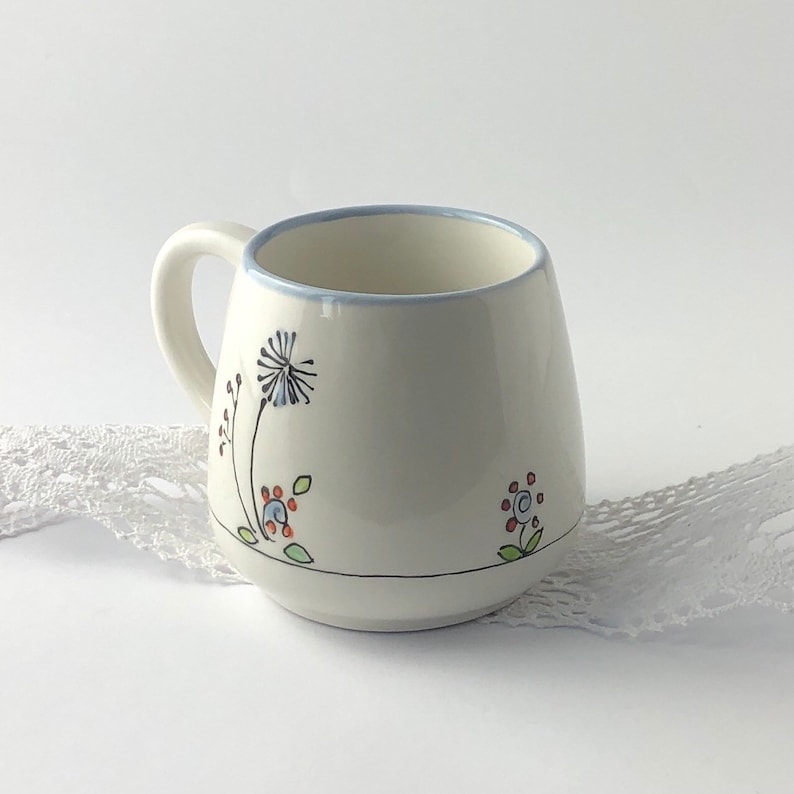 May include: White ceramic mug with a blue rim and hand-painted floral designs in shades of blue, red, and green. The design includes a dandelion, two small flowers, and a few leaves.