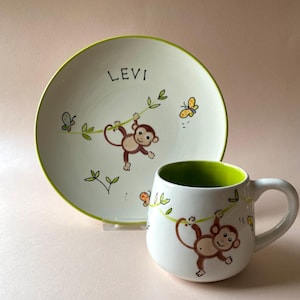 Pode incluir: Um prato de cerâmica branco com uma borda verde e um desenho de macaco. O prato tem o nome "Levi" escrito nele. Uma caneca de cerâmica branca combinando com uma borda verde e um desenho de macaco.