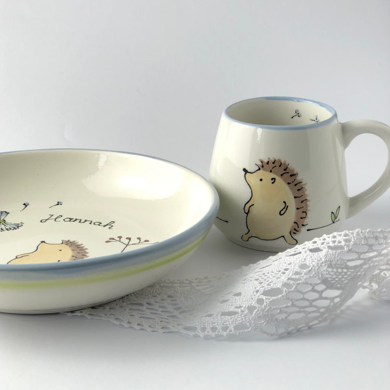 May include: A white ceramic children's dish and mug set with a hedgehog design. The dish has a blue and green rim and the mug has a blue rim. The dish is personalized with the name "Hannah".