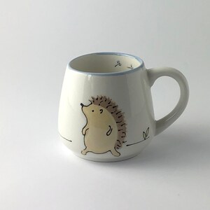 May include: White ceramic mug with a hand-painted illustration of a brown hedgehog walking on a white background. The mug has a blue rim and a handle.