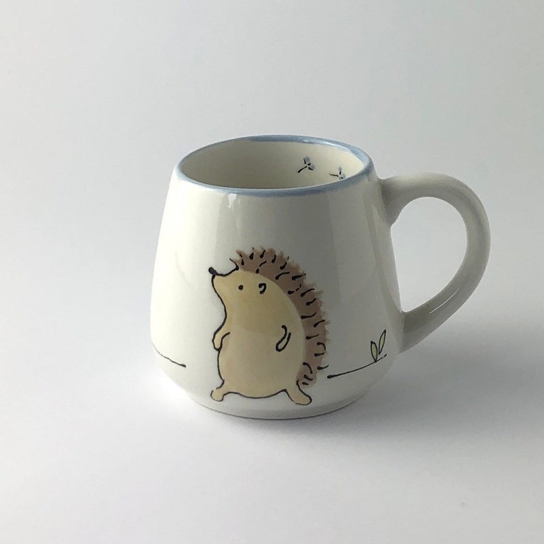 May include: White ceramic mug with a hand-painted illustration of a brown hedgehog walking on a white background. The mug has a blue rim and a handle.