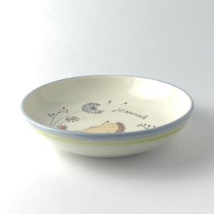 May include: A white ceramic bowl with a blue and green rim. The bowl has a hand-painted design of a hedgehog blowing on a dandelion with the name "Hannah" written on the bowl.