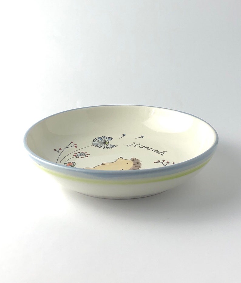 May include: A white ceramic bowl with a blue and green rim. The bowl has a hand-painted design of a hedgehog blowing on a dandelion with the name "Hannah" written on the bowl.