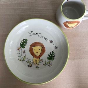 Pode incluir: Uma tigela de cerâmica branca com uma borda verde, apresentando uma ilustração pintada à mão de um leão, plantas da selva e flores. A tigela é personalizada com o nome "Luon" e a data "01.07.2020".