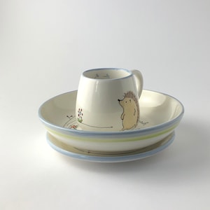 May include: A white ceramic children's dish set with a blue rim and a green stripe. The set includes a bowl and a small mug with a hedgehog illustration.
