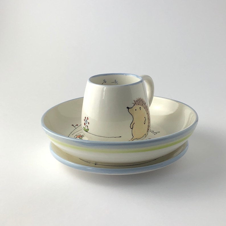 May include: A white ceramic children's dish set with a blue rim and a green stripe. The set includes a bowl and a small mug with a hedgehog illustration.