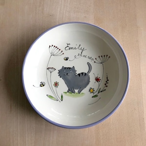 Può includere: Una ciotola in ceramica bianca con un bordo viola, con un design dipinto a mano di un gatto grigio, fiori e api. La ciotola è personalizzata con il nome "Emily" e la data "27.06.2020".