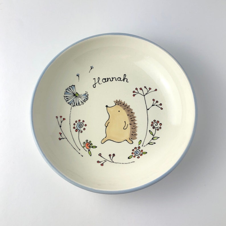 May include: A white ceramic bowl with a blue rim, featuring a hand-painted design of a hedgehog walking through a field of flowers. The bowl is personalized with the name "Hannah".