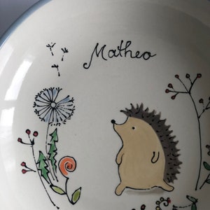 May include: A white ceramic bowl with a hand-painted design featuring a hedgehog, dandelions, and a flower. The bowl is personalized with the name "Matheo".