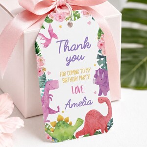 K&ouml;nnte beinhalten: Eine wei&szlig;e Geschenkbox mit einem hellrosa Satinband und einem rechteckigen Papieranh&auml;nger, der mit Aquarellillustrationen von Dinosauriern, rosa Blumen und gr&uuml;nen Bl&auml;ttern verziert ist. Der Text lautet Thank you FOR COMING TO MY BIRTHDAY PARTY! Love. Amelia.