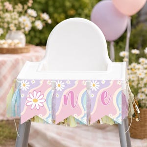 Op de afbeelding: Een witte kinderstoel versierd met een vlaggenlijn met een pastelroze, paars en blauw golvend patroon met witte madeliefjes. De banner toont de letters O, N en E op drie afzonderlijke vlaggetjes, geaccentueerd met hangende kwastjes.