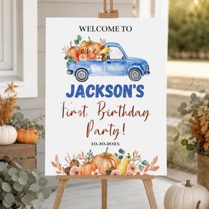 May include: A white rectangular sign on a wooden easel featuring a blue truck illustration, orange pumpkins, and text reading WELCOME TO JACKSON'S First Birthday Party! 10.10.202X. The design includes watercolour-style floral and pumpkin accents.