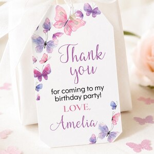May include: A white rectangular gift tag with clipped corners featuring watercolor butterflies in shades of pink and purple. The text reads Thank you for coming to my birthday party! LOVE, Amelia. The tag is tied with a white ribbon.