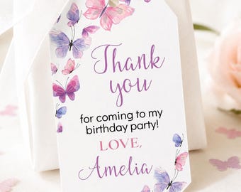 May include: A white rectangular gift tag with clipped corners featuring watercolor butterflies in shades of pink and purple. The text reads Thank you for coming to my birthday party! LOVE, Amelia. The tag is tied with a white ribbon.