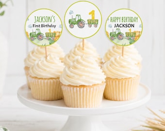 May include: Three cupcakes topped with circular paper toppers featuring watercolor green tractors, yellow number ones, and text reading Jackson's First Birthday and Happy Birthday Jackson, all displayed on a white pedestal stand.