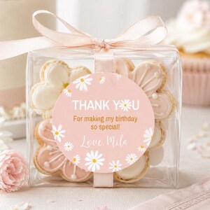 May include: A clear square gift box containing flower-shaped cookies, tied with a pale pink ribbon. A round sticker on the front features a pink background with white daisy illustrations and the text: THANK YOU For making my birthday so special! Love, Mila.