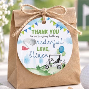 May include: A round paper gift tag tied with twine to a brown paper bag. The tag features a watercolor golf theme with a golf cart, balloons, and bunting. Text reads: THANK YOU for making my birthday onederful LOVE, Oliver, with a golf flag graphic.