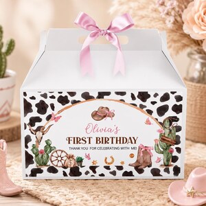 May include: A white cardboard gable box with a pink satin ribbon bow on top. The box features a black and white cow print pattern and a central graphic with a brown cowboy hat, cacti, a skull, a wagon wheel, and the text Olivia's FIRST BIRTHDAY.