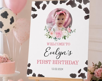 May include: A rectangular welcome sign on a wooden easel featuring a cow print border, a circular photo of a baby wearing a pink headband, and a floral arrangement. The text reads: WELCOME TO Evelyn's FIRST BIRTHDAY and the date 12.02.202X.