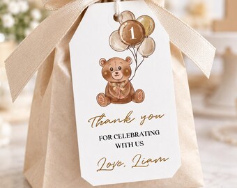 May include: A light brown paper favour bag tied with a beige satin ribbon and a white rectangular tag. The tag features a brown teddy bear holding balloons with the number 1, and the text: Thank you FOR CELEBRATING WITH US Love, Liam.