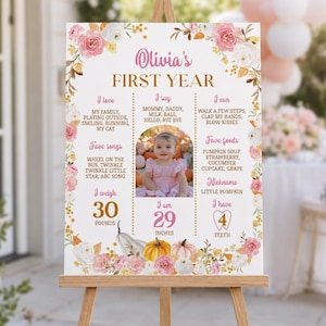May include: A white milestone birthday sign for Olivia's first year, decorated with pink and white roses and orange pumpkins. It features text detailing favorite songs, foods, and milestones, along with a central photo of a baby in a pink dress.