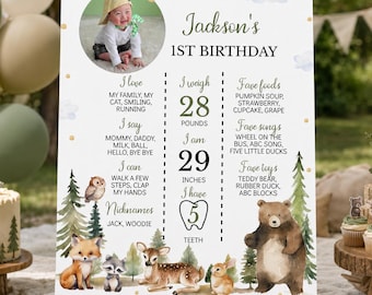 May include: A white birthday milestone sign for Jackson's 1st birthday featuring a circular photo of a child, watercolor forest animals, and text listing favorites, 28 pounds, 29 inches, and 5 teeth, displayed on a wooden easel stand.