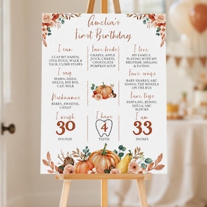 May include: A white milestone birthday sign for Amelia featuring orange pumpkin illustrations, floral accents, and text detailing milestones like 30 pounds, 4 teeth, and 33 inches, displayed on a wooden easel with a fall-themed color palette.