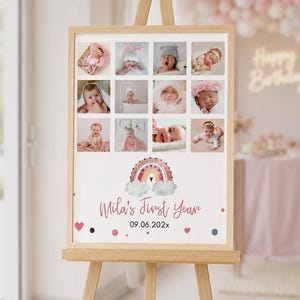 May include: A rectangular sign on a wooden easel featuring a twelve-photo collage of a baby. Below the photos is a watercolour rainbow illustration with clouds and the text Mila's First Year 09.06.202x, accented by small pink hearts and dark dots.