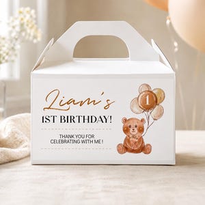May include: A white cardboard gable box featuring a brown teddy bear illustration holding a cluster of balloons with the number 1. The text reads: Liam's 1ST BIRTHDAY! THANK YOU FOR CELEBRATING WITH ME! The design uses brown and black lettering.