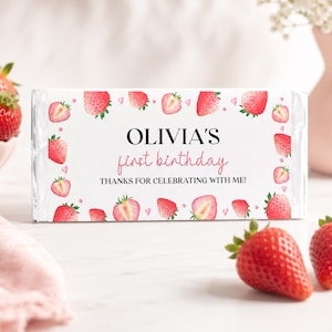 May include: A rectangular candy bar wrapper with a white background featuring a pattern of red strawberries and small pink hearts. The text reads OLIVIA'S first birthday THANKS FOR CELEBRATING WITH ME! in black and pink serif and script fonts.