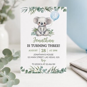 May include: A rectangular birthday invitation card featuring a watercolor illustration of a koala holding a light blue balloon with the number 3. The card includes green eucalyptus foliage at the top and bottom, with text detailing Jonathan's third birthday party information.