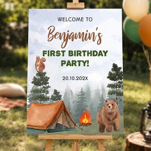 Op de afbeelding: Een rechthoekig verjaardagsbord op een houten ezel met een bossc&egrave;ne, een bruine tent, een kampvuur, een eekhoorn en een beer. De tekst luidt: WELCOME TO Benjamin's FIRST BIRTHDAY PARTY! 20.10.202X. De achtergrond toont bomen in aquarel-stijl.