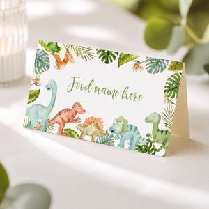 Może przedstawiać: Złożona biała kartka z akwarelową ilustracją r&oacute;żnych dinozaur&oacute;w i tropikalnych zielonych liści. Tekst "Food name here" jest wydrukowany zieloną kursywą na środku kartki, w otoczeniu kolorowych dinozaur&oacute;w.