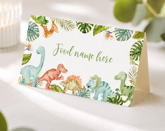 May include: A folded white card featuring a watercolor illustration of various dinosaurs and tropical green leaves. The text "Food name here" is printed in a green cursive script in the center of the card, surrounded by colorful dinosaurs.