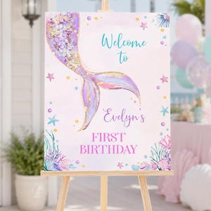 May include: A rectangular sign on a wooden easel featuring a watercolor mermaid tail in shades of purple, pink, and gold. The text reads Welcome to Evelyn's FIRST BIRTHDAY in teal and purple script and block lettering, surrounded by sea life.