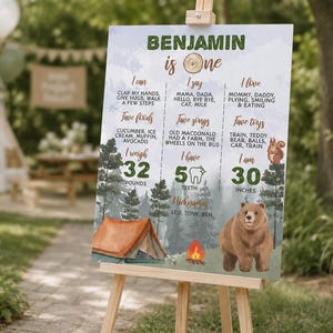 May include: A rectangular milestone birthday sign for a one-year-old named Benjamin, featuring a forest theme with a brown bear, a tent, and a campfire. The text lists milestones including weight in pounds, height in inches, and favorite things.