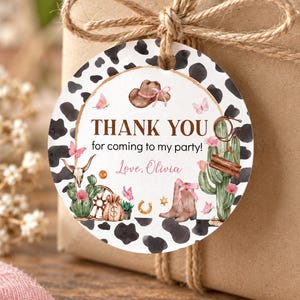 K&ouml;nnte beinhalten: Ein rundes Geschenkanh&auml;nger mit schwarz-wei&szlig;em Kuhfleckenmuster am Rand, verziert mit Aquarellillustrationen von Cowboyhut, Kaktus, Stiefeln und Schmetterlingen. Der Text lautet: THANK YOU for coming to my party! Love, Olivia, in Braun und Rosa.