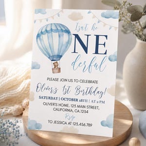 Pu&ograve; includere: Un biglietto d'invito di compleanno rettangolare bianco con una mongolfiera blu ad acquerello e un piccolo orso bruno nel cesto. Il testo recita Isn't he ONEderful, PLEASE JOIN US TO CELEBRATE Oliver's 1st Birthday e i dettagli dell'evento.