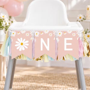 May include: A high chair banner featuring three light pink pennants with white daisy patterns and the letters O, N, and E in white. The banner is decorated with hanging tassels in shades of light pink, yellow, and metallic gold.