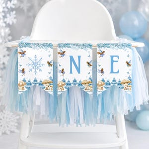 May include: A high chair banner featuring three white pennants with blue snowflake, fairy, and mushroom illustrations. The letters O, N, and E are printed in blue on each pennant. The banner is decorated with light blue and white hanging fringe.